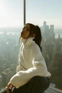 A woman wearing an oversized cream turtleneck knit sweater, sitting by a window with a blurred city skyline in the background.