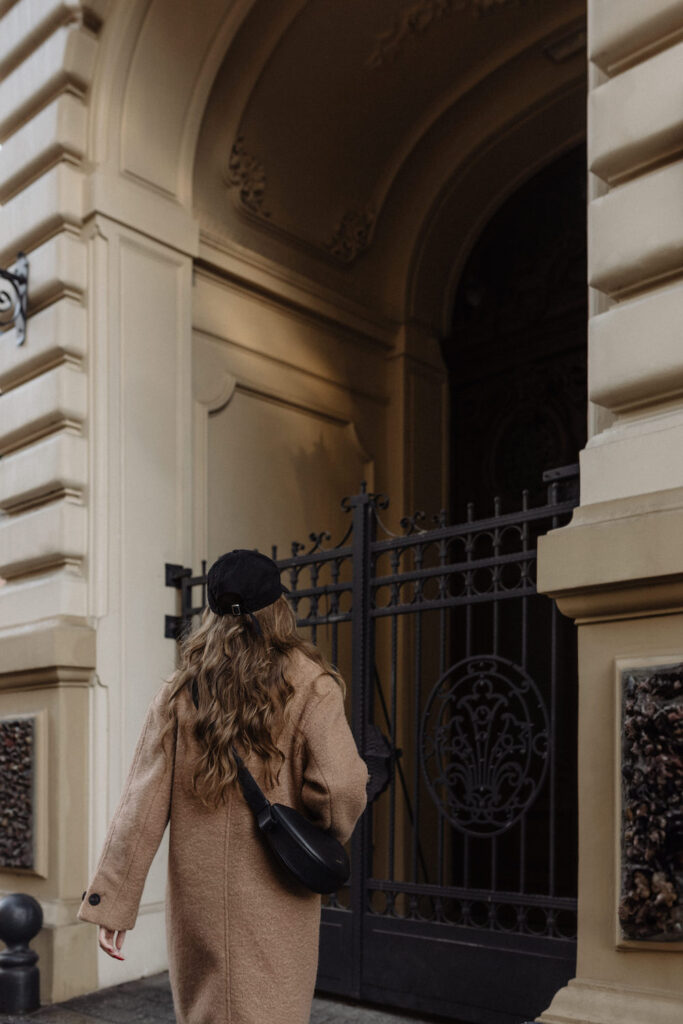 A modern tailored style woman wearing a black cap, a long brown coat and a black bag draped across her shoulder.
