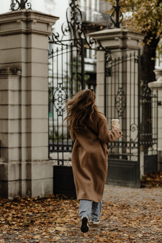 A woman walking to a stone building with black gates wearing an oversized brown wool coat and light-wash jeans carrying a cup of coffee in her right hand for The Capsule Wardrobe Essentials Edit.