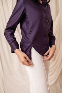 The curated edit brings you a close-up of a deep purple satin button-down blouse with side-button detailing paired with white tailored trousers for our modern tailored style.