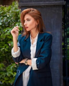 A woman wearing a teal-blue blazer with rolled up sleeves, layered over a white shirt, standing by a pillar.
