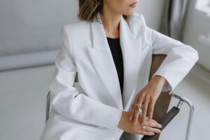 A woman sitting in a chair wearing a white blazer layered over a black top.