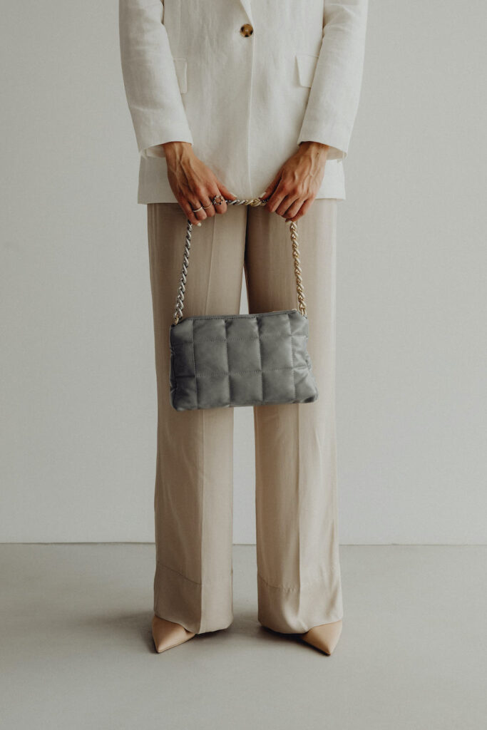 A woman standing wearing a white blazer and beige trousers holding a silver-grey quilted bag form the Spring Work Wear Edit.