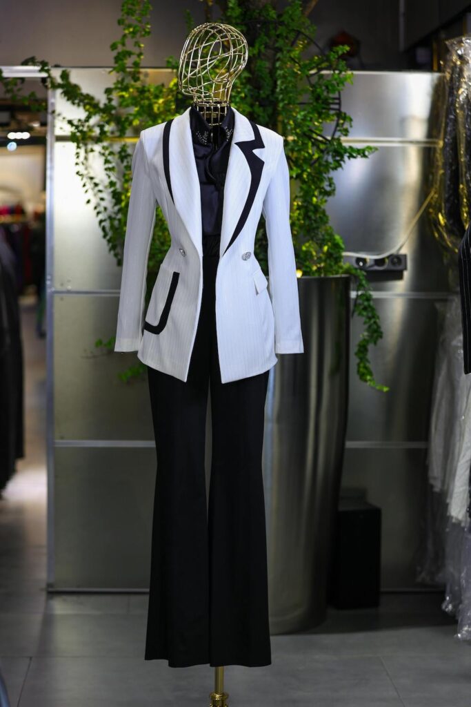 Modern tailored style white blazer with black contrast piping shown with wide-leg black trousers on a mannequin.