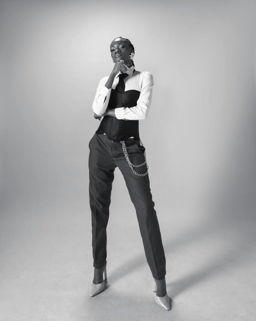 For the Capsule Wardrobe Essentials, we have a woman standing in a thoughtful pose wearing a white button down shirt with a black tie, a black corset layered over the shirt and charcoal tailored pants with silver chains draped on the left pocket.