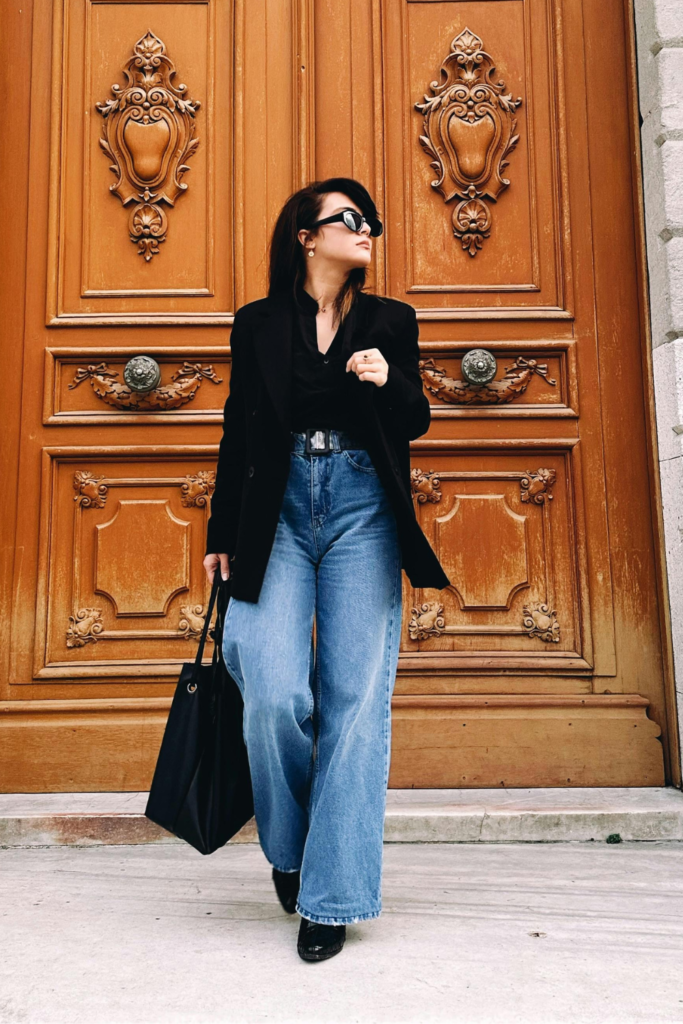 Woman wearing a structured black blazer and wide-leg denim jeans in front of ornate wooden doors.