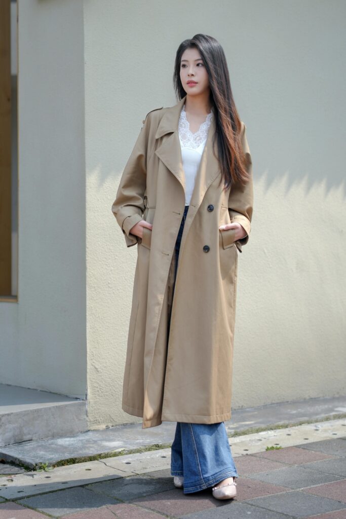 A woman standing on a city sidewalk next to a building. She is wearing a long, loose-fitting tan trench coat over a white lace-trimmed top and wide-leg blue flare jeans.