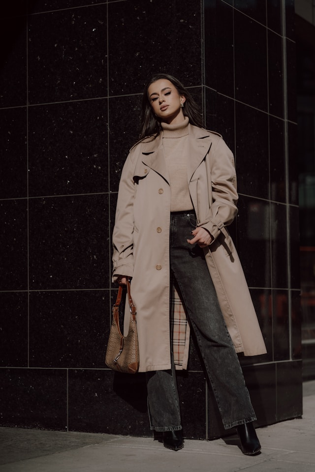 A woman leaning against a dark, glossy tiled building wall on a city street. She is wearing an open, relaxed beige trench coat over a tonal beige turtleneck, dark grey wide-leg jeans, and black boots, holding a patterned shoulder bag.