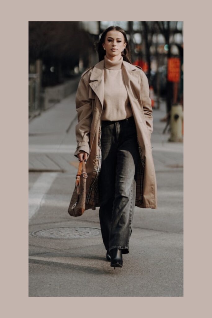 A woman walking forward on a city street wearing an oversized beige trench coat, a matching cream turtleneck knit, dark wash jeans, and black boots. She carries a patterned tote bag in one hand, framed against a soft beige border.
