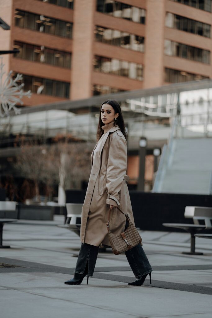 Side profile of a woman walking in a city plaza wearing a beige trench coat with a belted back, dark wide-leg jeans, black high-heeled boots, and carrying a brown patterned shoulder bag.
