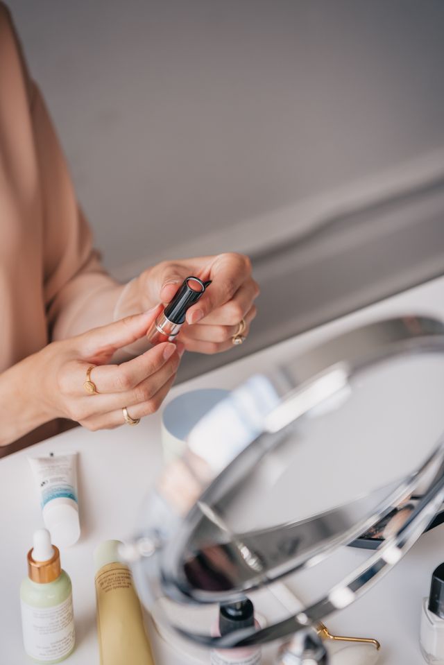 Close-up photograph focusing on two hands wearing gold rings holding an open tube of coral-pink lipstick, with a bright white makeup table, skincare bottles, and a round silver vanity mirror in the softly blurred foreground.