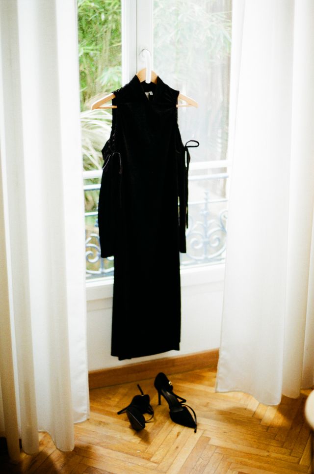 A minimalist bedroom view showing a black, high-neck, long-sleeved dress with unique shoulder details hanging on a wooden hanger by a window with white curtains. Below the dress, on the wooden floor, are a pair of matching black stiletto heels.