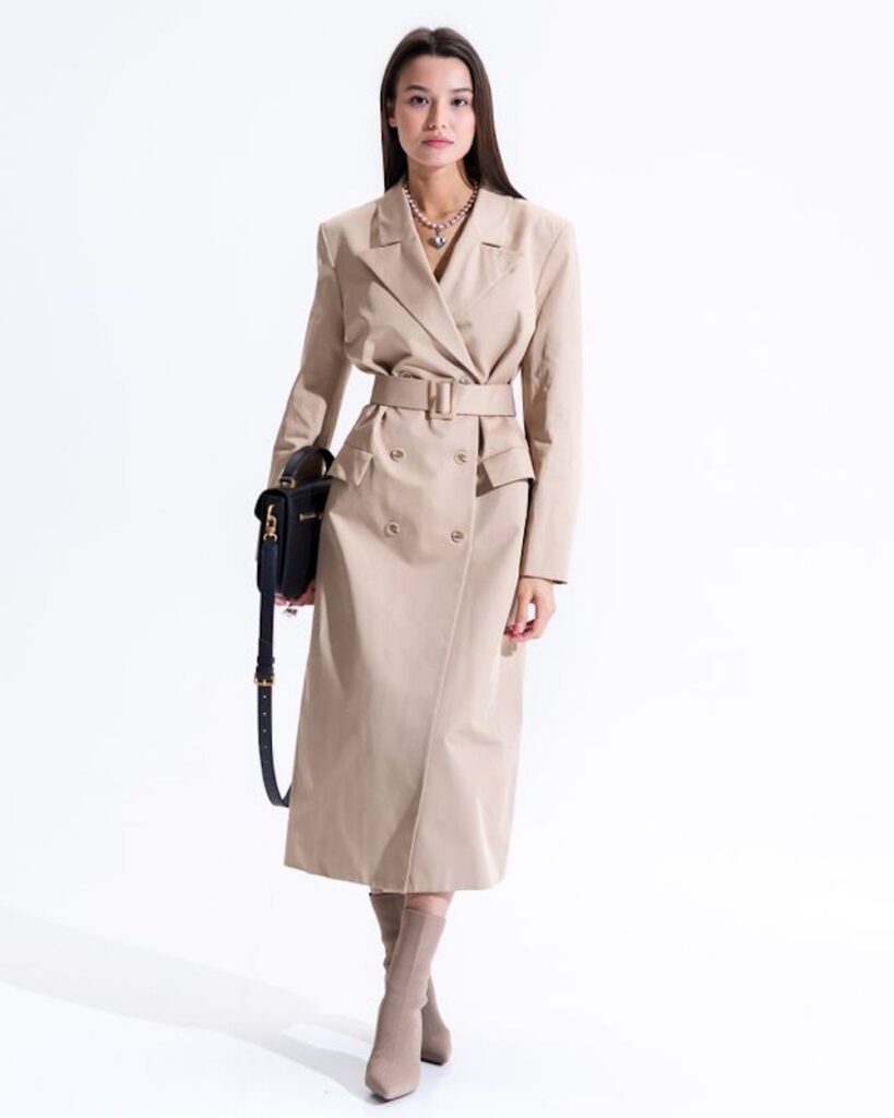 A woman posing against a white background wearing a tailored, double-breasted beige trench coat fully buttoned and tightly belted at the waist like a dress. She is wearing matching beige sock boots, a statement chain necklace, and holding a structured black leather handbag.