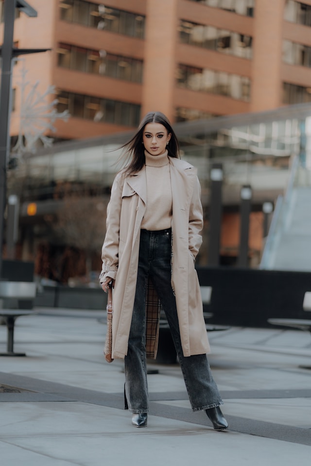 In our trench coat for women edit, showcases a woman standing in an urban plaza wearing a classic beige trench coat draped over a light beige turtleneck sweater and dark grey wide-leg denim jeans with split hems. She is wearing black heeled ankle boots and holding a patterned brown shoulder bag.
