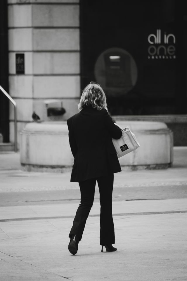 A black and white candid photograph taken from behind showing a woman walking down a city street wearing a tailored blazer, flared trousers, heels, and carrying a structured canvas tote bag.