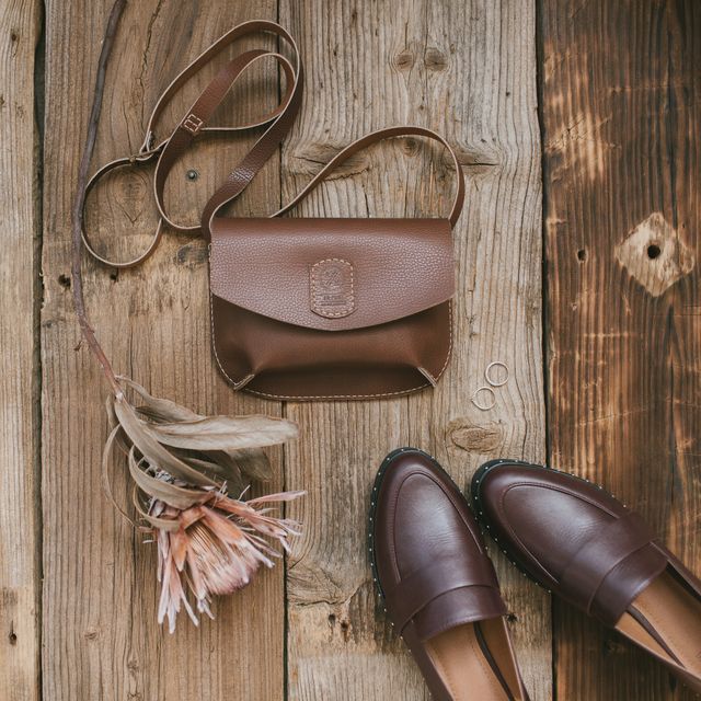 An overhead flat-lay on a rustic wooden surface featuring a structured brown leather handbag, a pair of classic leather loafers, a dried protea flower, and two simple silver rings.