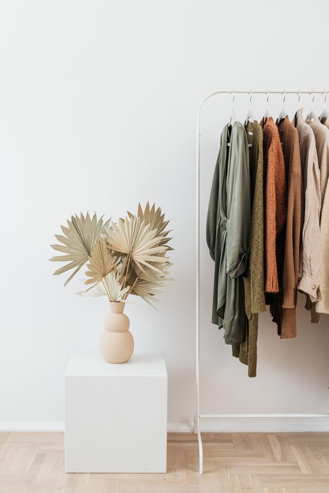 A minimal white clothing rack holding a curated selection of women's autumn-toned sweaters and dresses next to a beige ceramic vase with dried palm leaves.