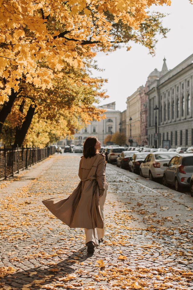 Rear view of a woman walking down a city street paved with cobblestones under golden autumn trees, her timeless camel trench coat billowing behind her to show modern classic style in motion. 
