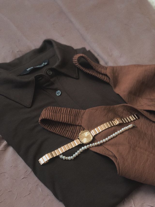 Elegant flat lay of a dark brown polo shirt paired with a brown silk garment, a classic gold square-faced watch, and a delicate pearl bracelet, representing a curated capsule wardrobe layout formula. 