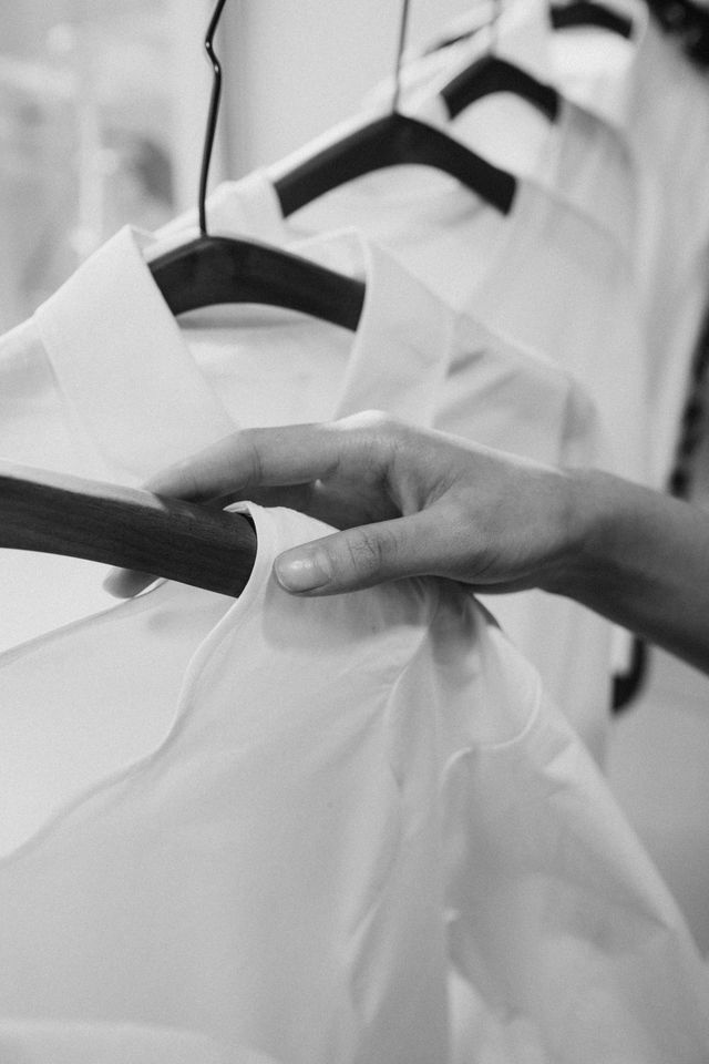 Black and white close-up of a woman's hand gently selecting a crisp white button-down shirt from a clothing rail, emphasising intentional wardrobe care and classic styling. 