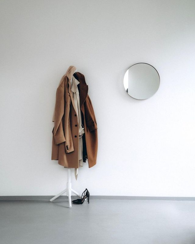 Minimalist white room for dressing with intention featuring a modern coat rack holding a classic camel trench coat and tailored outerwear next to a round wall mirror, showcasing timeless capsule wardrobe essentials. 