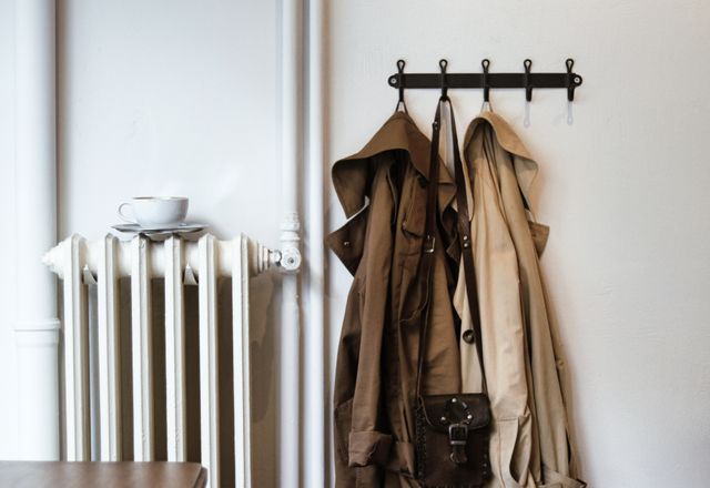 A capsule wardrobe mindset these two classic trench coats and a small leather crossbody bag hanging on a vintage wall rack next to a white radiator with a coffee cup on top.
