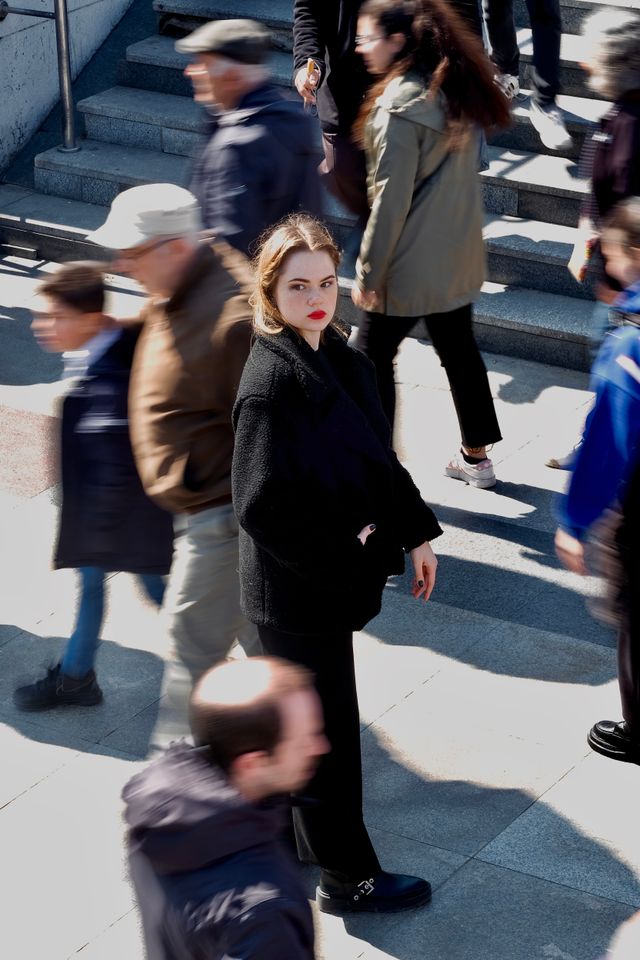 Overhead view of a woman in a black coat with red lipstick standing still amidst a blurred, fast-moving crowd on outdoor city steps.

