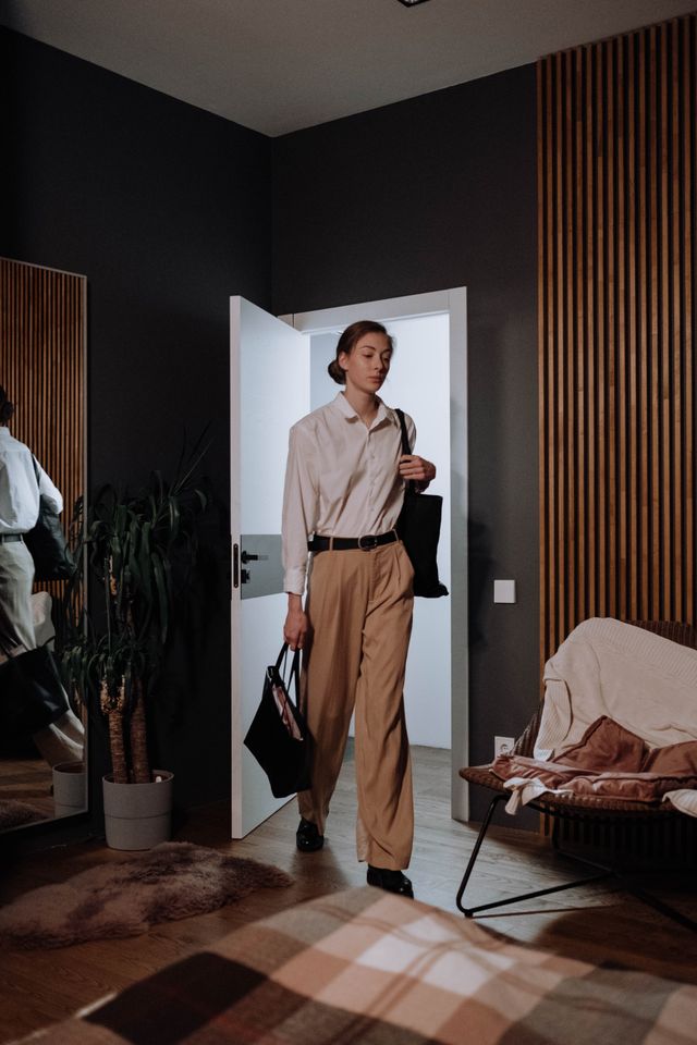 Professional woman wearing high-waisted beige trousers, a white button-down shirt, and black loafers carrying a tote bag through a modern interior doorway. 