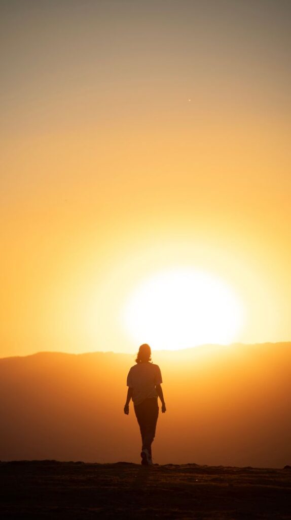Silhouette of a woman walking away toward a massive, bright glowing sun at sunset with mountains in the background.