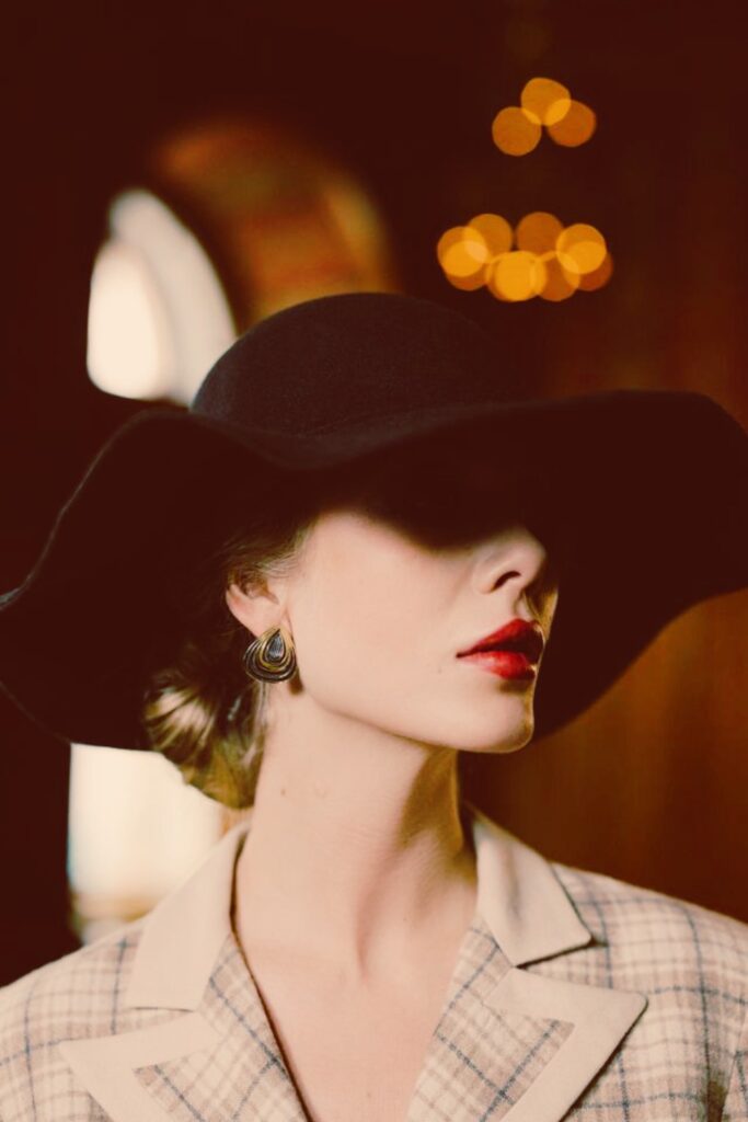  Close-up profile portrait of an elegant woman wearing a wide-brimmed black hat casting a shadow over her eyes, styled with red lipstick, a gold statement earring, and a plaid blazer.
