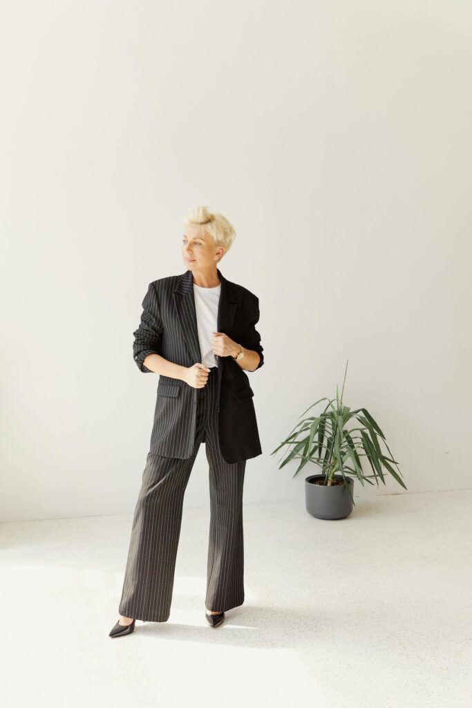 Woman with blonde crop haircut wearing an oversized black pinstripe suit, white t-shirt, and black pointed heels standing in a bright minimalist studio for our modern classic work formula.