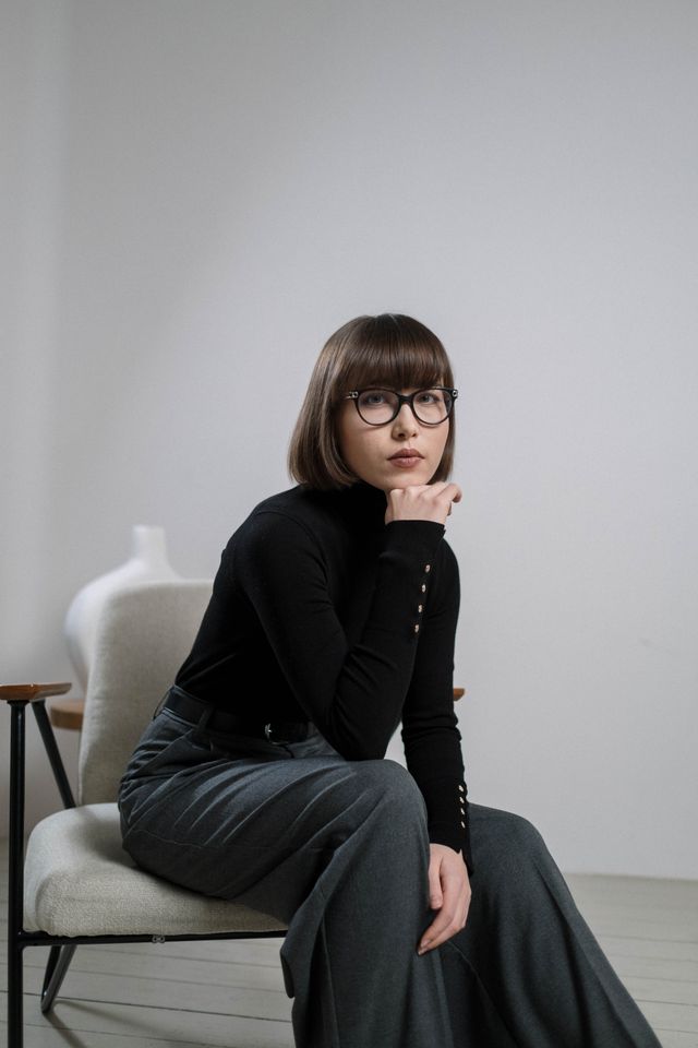 A chic woman with a bob haircut and glasses wearing a black turtleneck and grey tailored trousers, seated in a minimal interior space.
