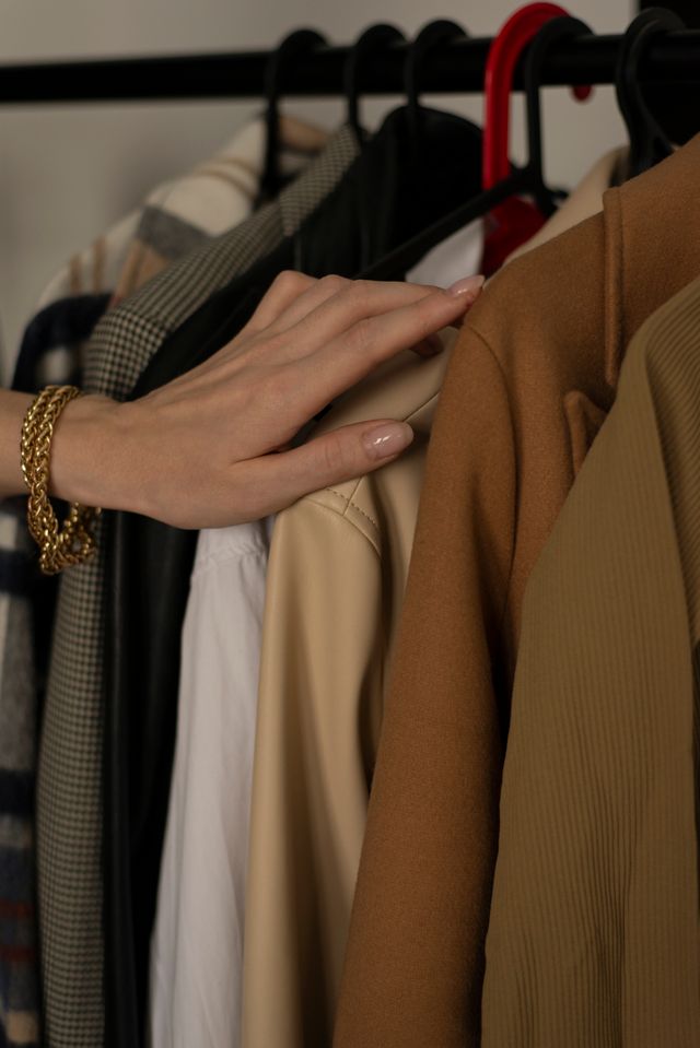Close-up of a woman's hand wearing a gold chain bracelet selecting a camel blazer from an organised clothing rack of classic women's workwear.