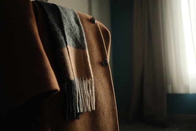 Close-up of a classic camel wool coat draped with a neutral patterned winter scarf near a sunlit window.