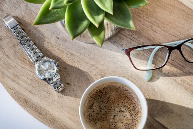 A flat-lay of the morning essentials featuring a silver wristwatch, eyeglasses, a cup of coffee, and a small green succulent on a wooden tabletop. 
