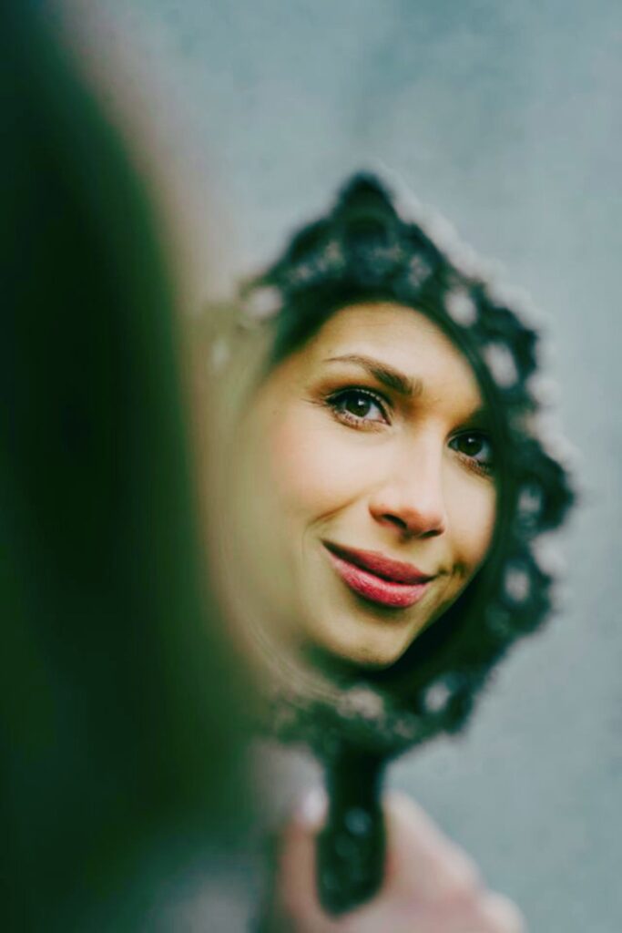 Close-up reflection of a woman with red lipstick smiling softly into an ornate, vintage hand mirror against a muted teal background.