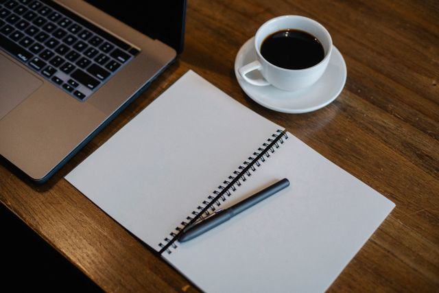  Overhead view of an open blank spiral notebook with a pen, sitting next to a white cup of black coffee and a laptop on a warm wooden desk