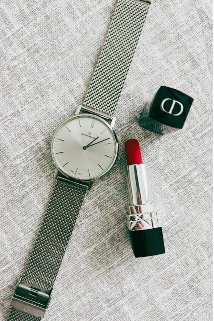 Flat-lay of a minimalist women's silver mesh strap watch next to an open tube of classic bold red lipstick on a textured neutral fabric background